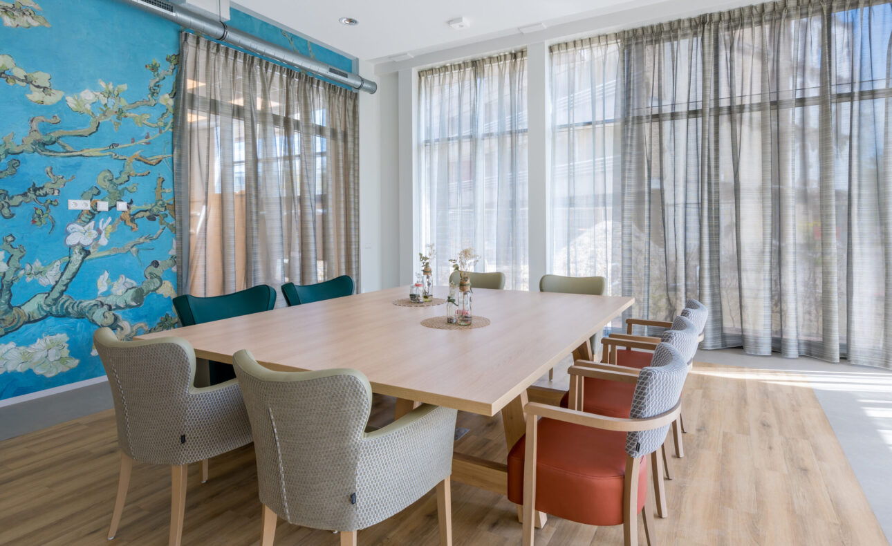 Op de foto is één van de zalen te zien die aangrenzend aan de brasserie is te vinden. De zaal heeft hoge ramen waarvoor lichtdoorlatende vitrage is gehangen. In het midden staat een grote houten tafel en daaromheen staan stoelen.