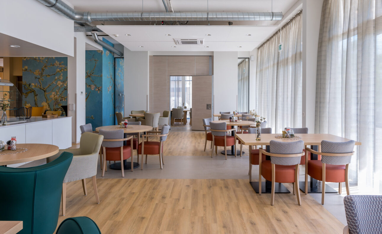 Op de foto is de brasserie van Bloesemhove te zien. Het is een ruime, lichte, hoge zaal waar overal zitjes te vinden zijn. Er staan oranje stoelen en er ligt een houten vloer.