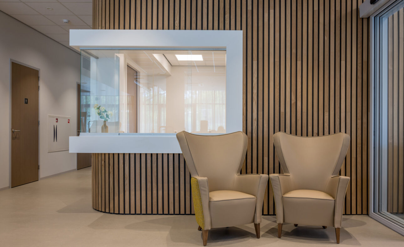 Op de foto is de receptie van Amandelhof te zien. Een strak afgewerkte balie met een sfeervolle houten lattenwand. Voor de receptie zijn twee fauteuils geplaatst.