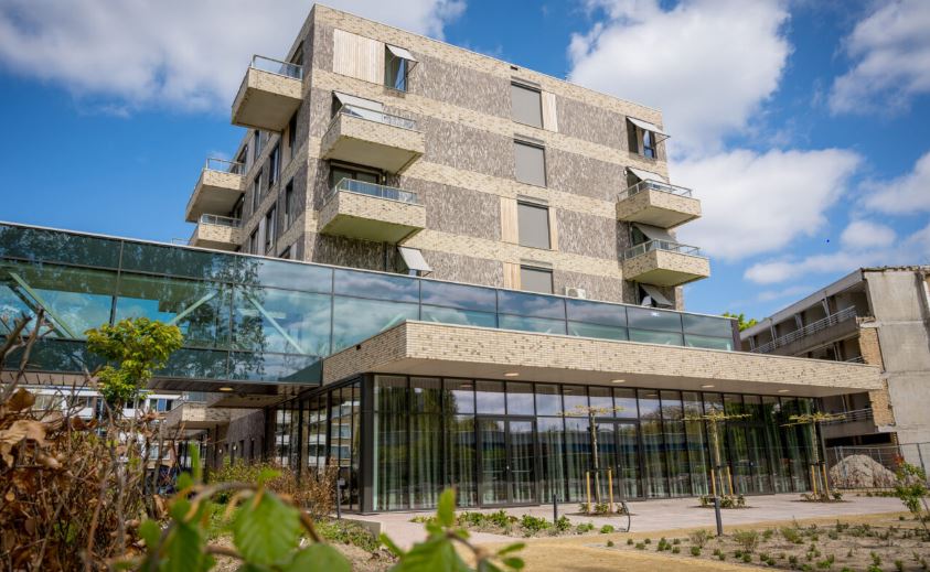 Buitenaanzicht van een modern gebouw met glazen gevel en balkons, omgeven door een tuin en blauwe lucht met wolken.