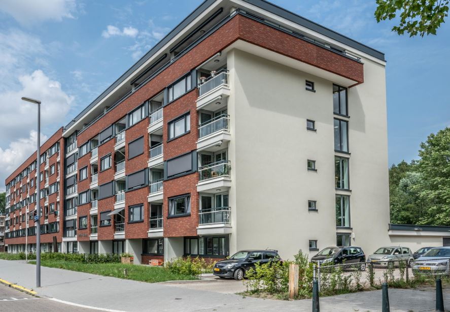 Zijkant van een modern appartementencomplex met rode bakstenen en balkons, geparkeerde auto's en een blauwe lucht op de achtergrond.