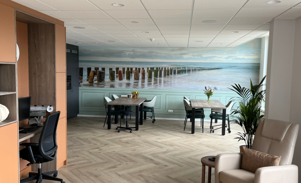 Moderne kantoorruimte met houten vloer, tafels en stoelen. Een grote muurschildering van een strand siert de achterwand.