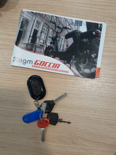 Sleutels met een blauwe en rode sleutelhanger liggen op een houten tafel naast een handleiding voor een AGM Goccia scooter.