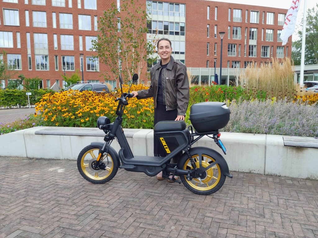 Persoon poseert met een zwarte elektrische scooter met gele accenten voor een gebouw, omringd door bloemen en planten.