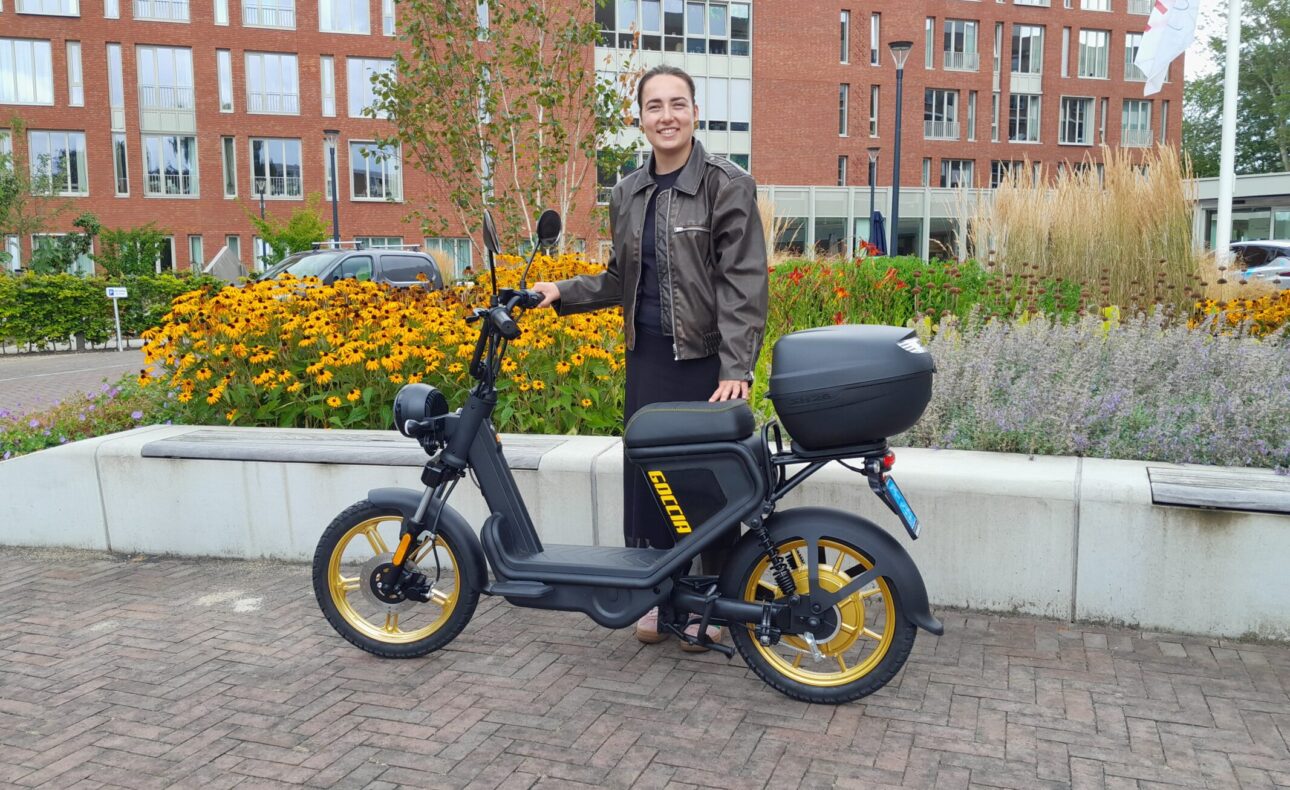 Persoon poseert met een zwarte elektrische scooter met gele accenten voor een gebouw, omringd door bloemen en planten.