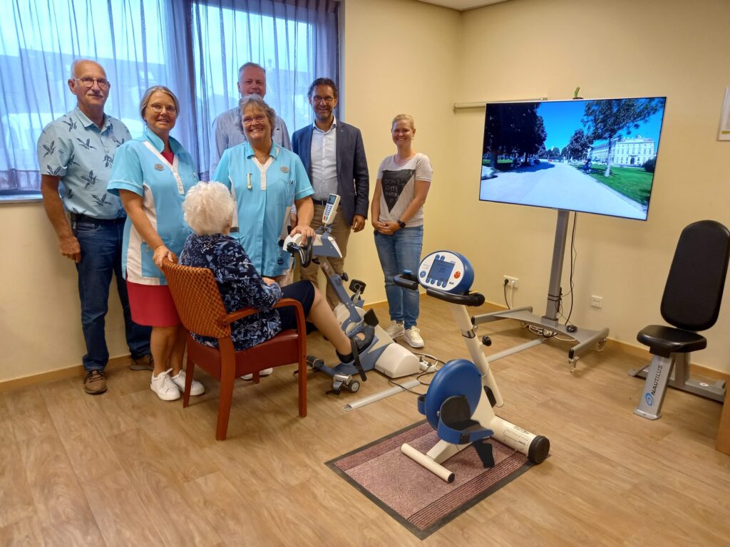 Groep mensen in een kamer met een oudere vrouw op een hometrainer. Een groot scherm toont een buitenomgeving.