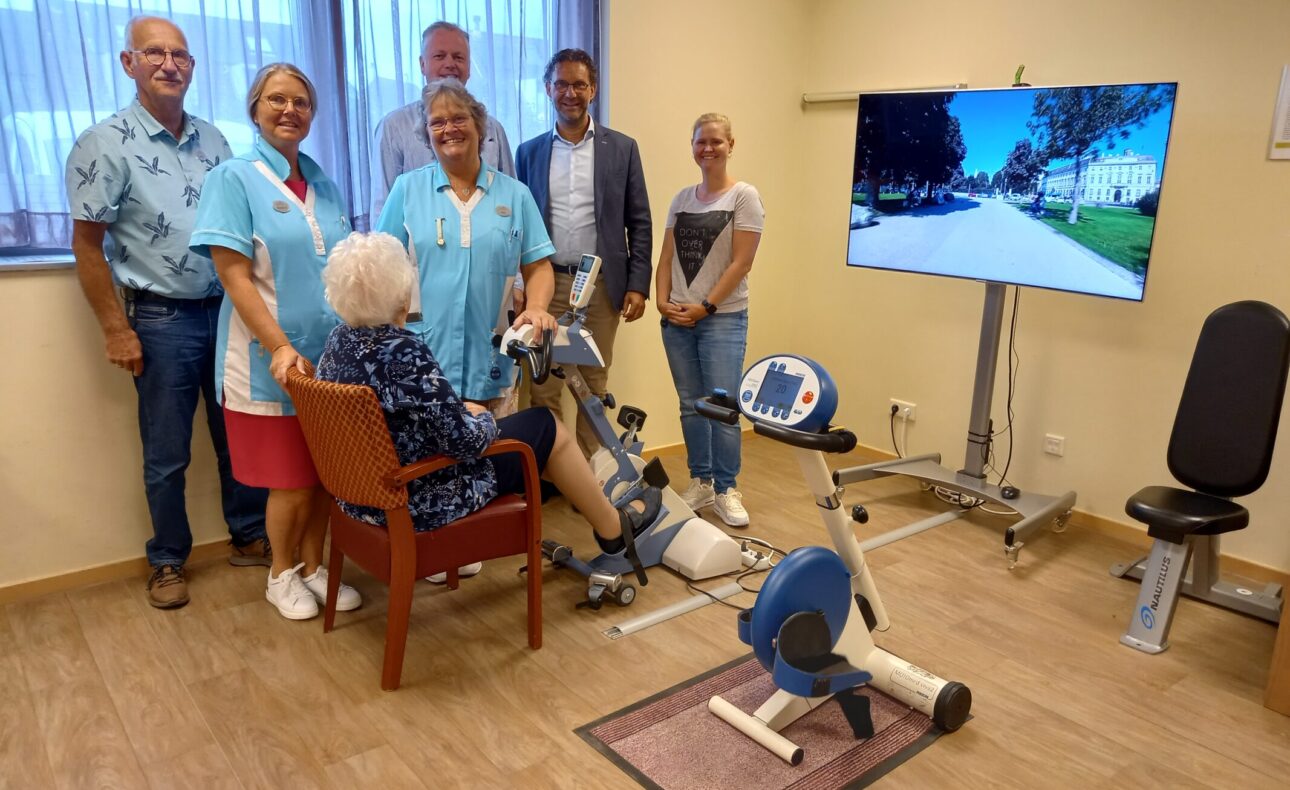 Groep mensen in een kamer met een oudere vrouw op een hometrainer. Een groot scherm toont een buitenomgeving.