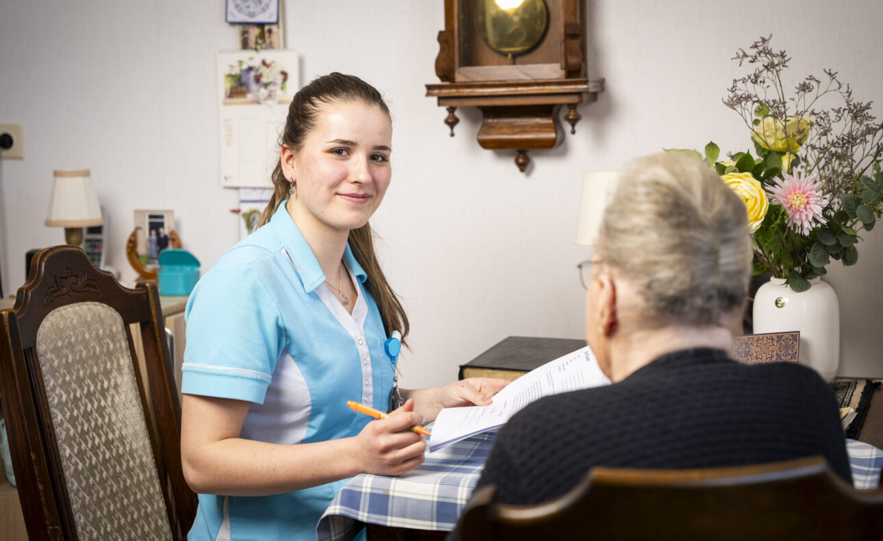 Een zorgverlener in een blauw uniform zit aan tafel met een oudere vrouw. Ze glimlacht en houdt een pen vast bij een notitieblok.