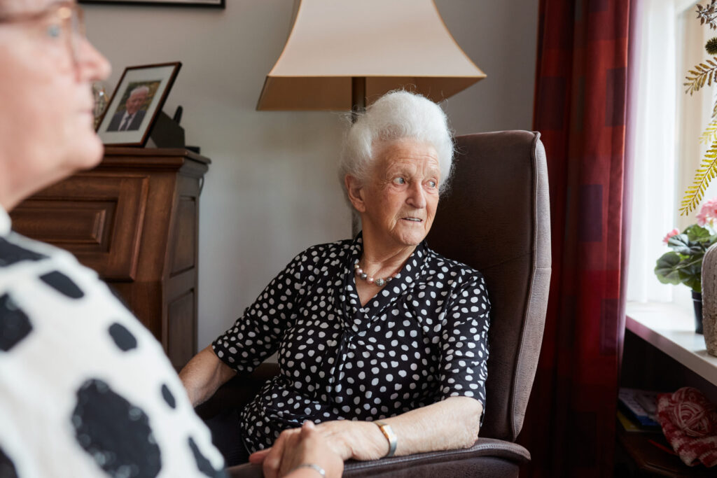 Een oudere vrouw met wit haar zit in een stoel bij een raam, gekleed in een zwart-wit gestippelde blouse, naast een lamp.