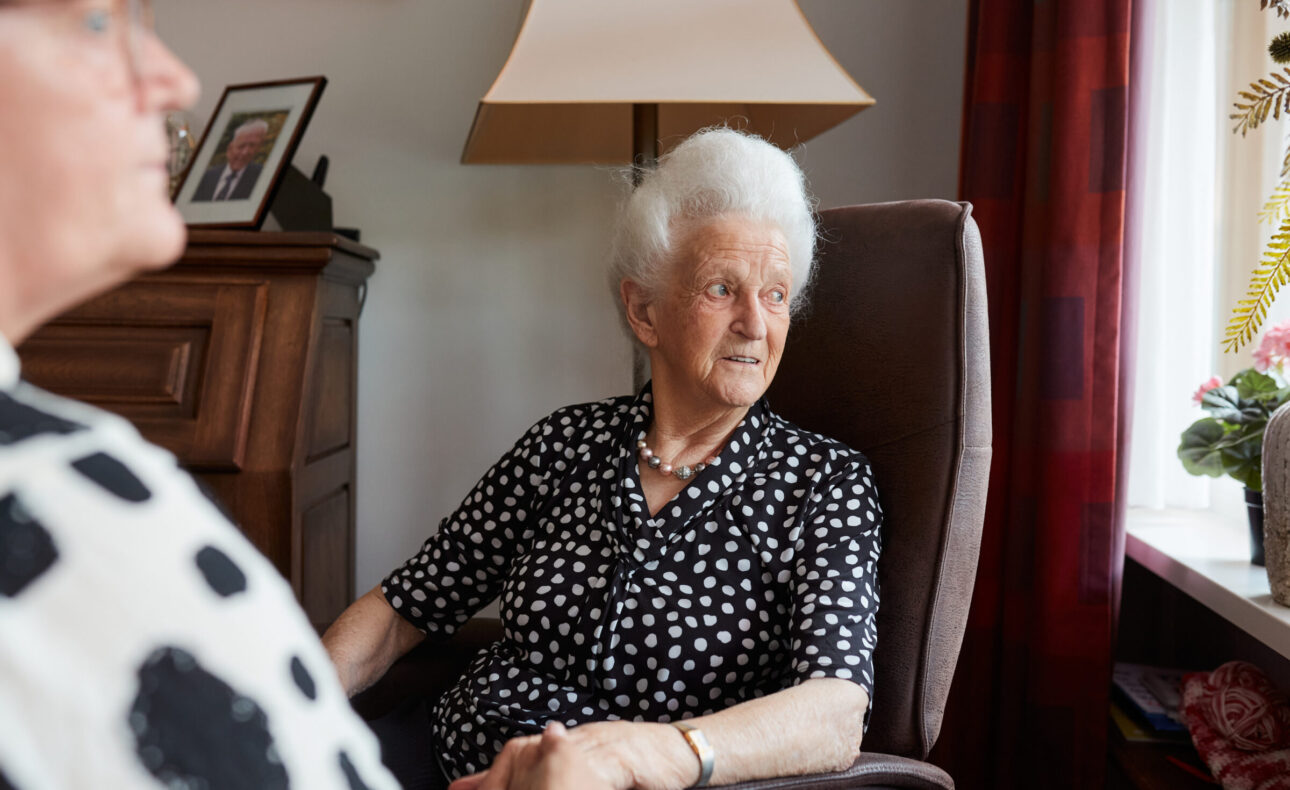 Een oudere vrouw met wit haar zit in een stoel bij een raam, gekleed in een zwart-wit gestippelde blouse, naast een lamp.
