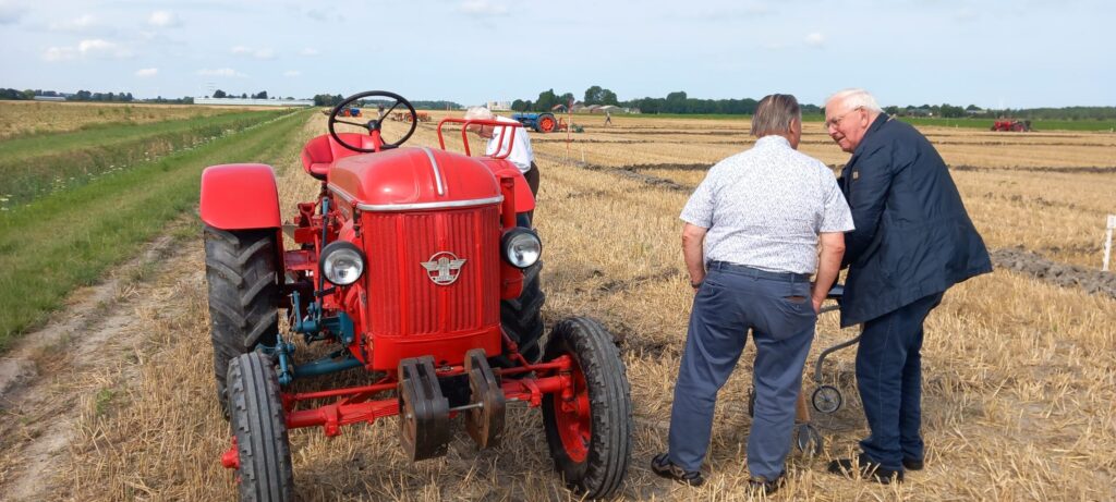 Twee mannen staan naast een rode tractor op een veld. Ze lijken in gesprek te zijn tijdens een ploegwedstrijd op het platteland.