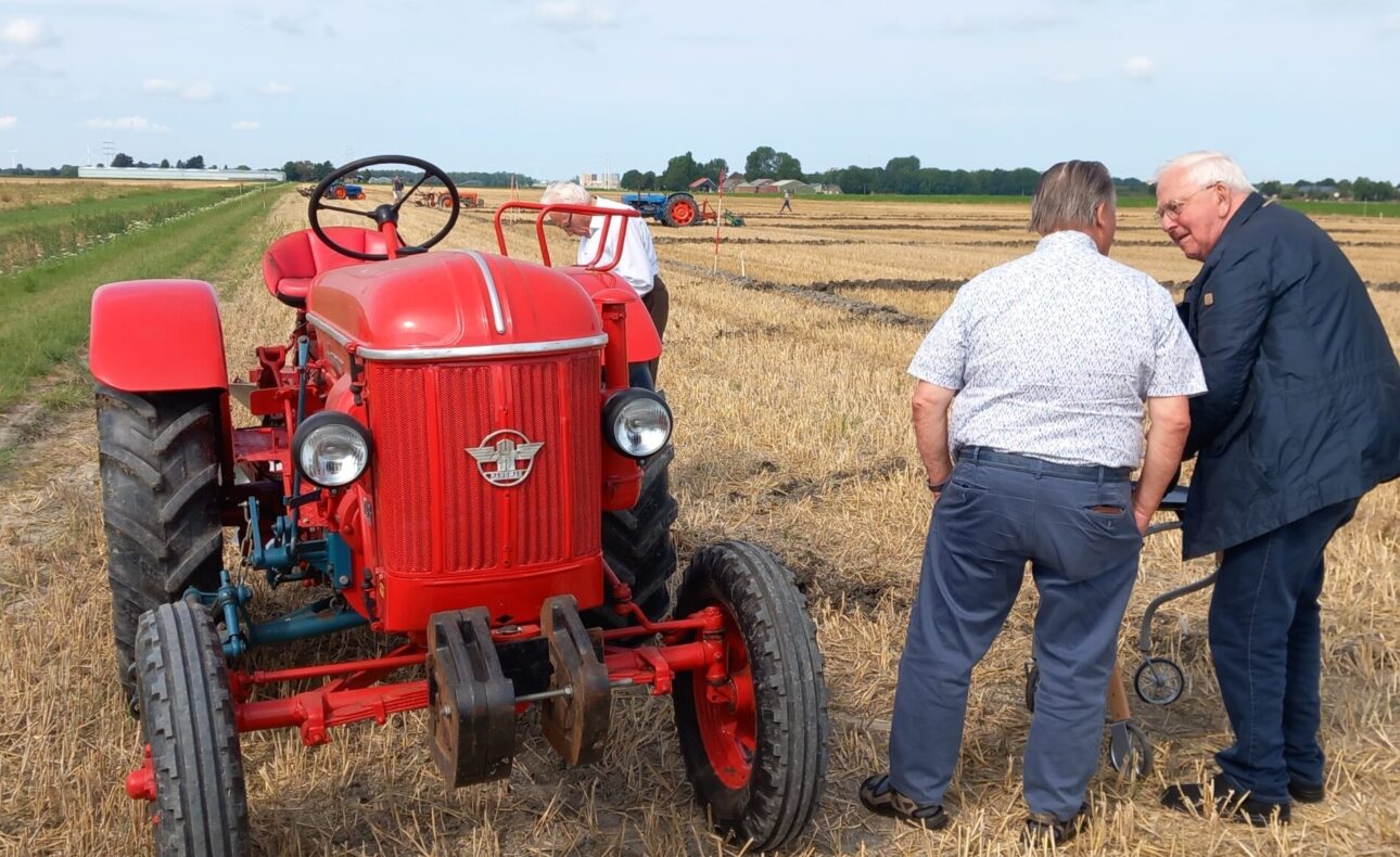 Twee mannen staan naast een rode tractor op een veld. Ze lijken in gesprek te zijn tijdens een ploegwedstrijd op het platteland.
