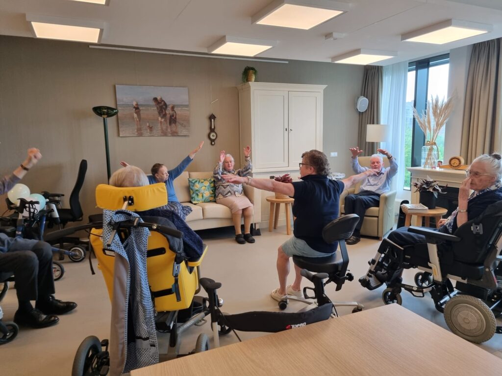 Groep ouderen in rolstoelen doet samen oefeningen in een lichte kamer met schilderij en grote ramen. Ze strekken hun armen uit.
