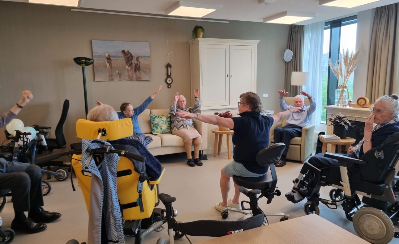 Groep ouderen in rolstoelen doet samen oefeningen in een lichte kamer met schilderij en grote ramen. Ze strekken hun armen uit.