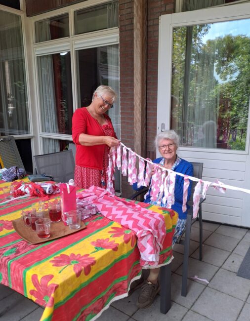 Twee vrouwen zitten buiten aan een tafel met kleurrijke tafelkleden, bezig met het maken van een stoffen slinger.