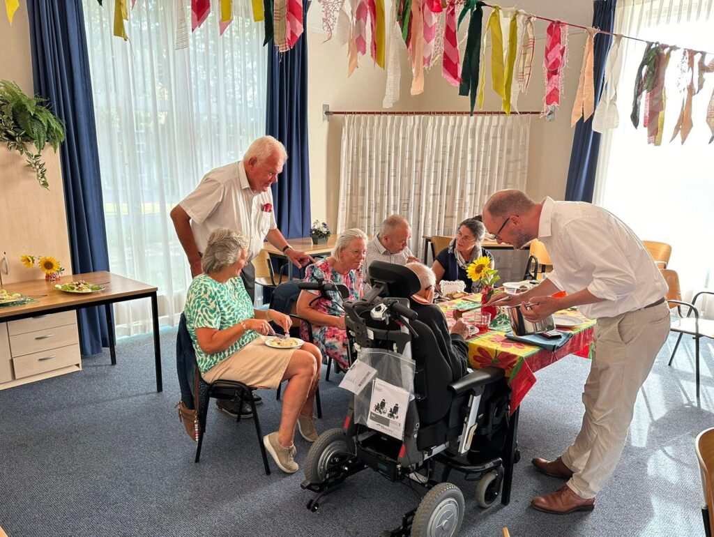 Een groep ouderen zit aan een tafel in een feestelijk versierde ruimte. Twee mannen staan erbij en praten met de groep.