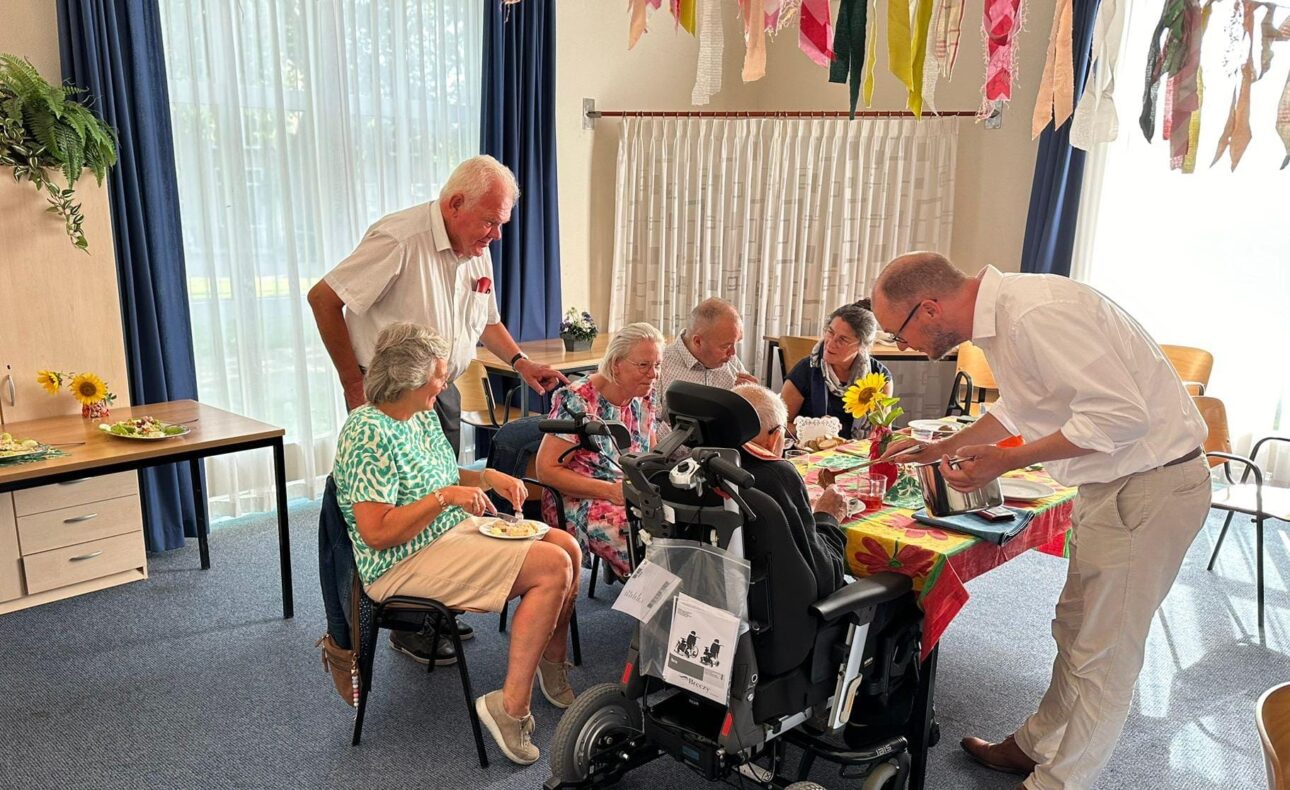Een groep ouderen zit aan een tafel in een feestelijk versierde ruimte. Twee mannen staan erbij en praten met de groep.
