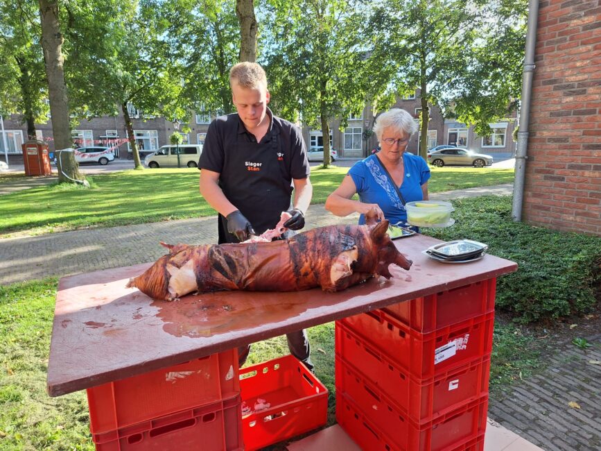 Twee mensen bereiden een geroosterd varken op een tafel buiten, omringd door bomen en gebouwen op een zonnige dag.