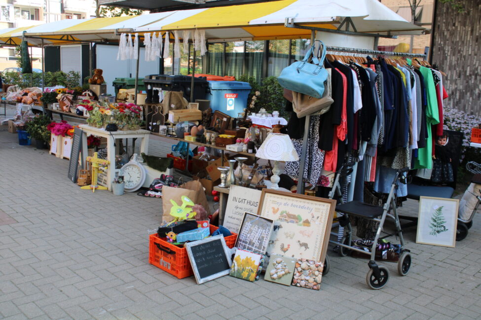 Kraampje op een markt met diverse tweedehands spullen, zoals kleding, boeken en decoraties, uitgestald onder een geel-witte luifel.