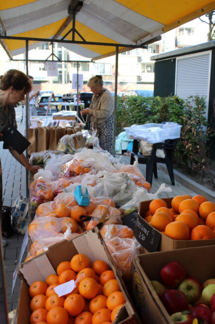 Een marktkraam met vers fruit en groenten, waaronder sinaasappels en appels. Twee mensen bekijken de producten onder een geel-wit gestreepte luifel.