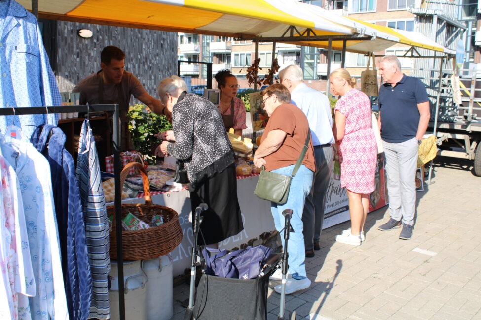 Mensen bekijken producten bij een kraam op een markt. Er hangen kledingstukken en er zijn manden met goederen zichtbaar.