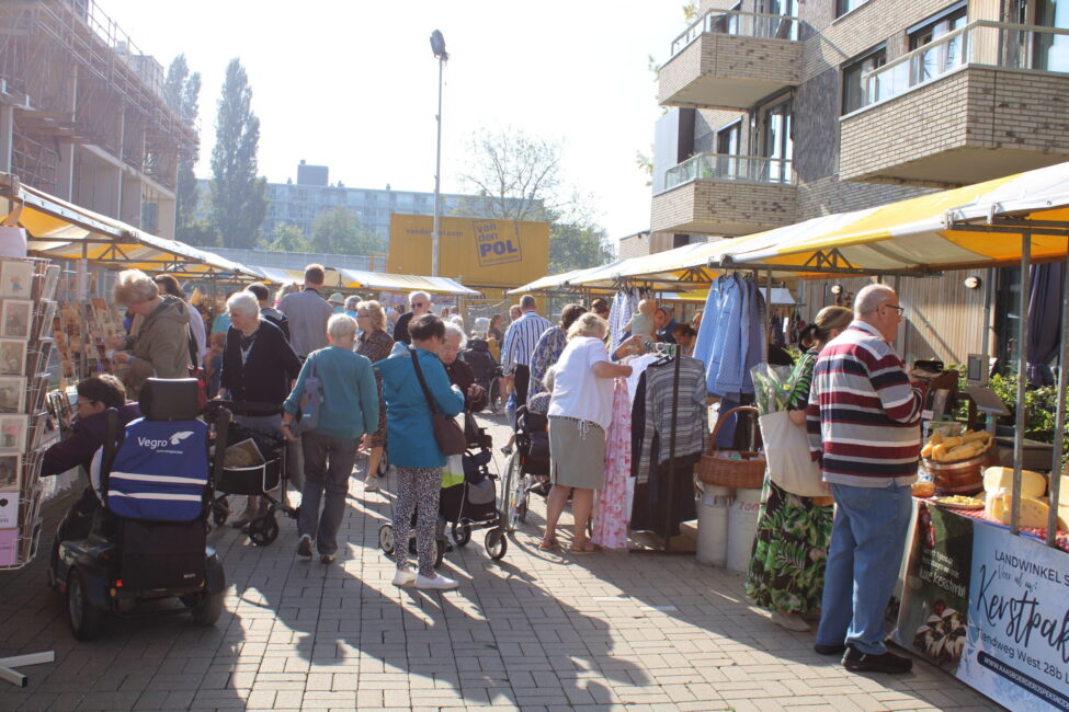 Een drukbezochte markt met mensen die langs kraampjes lopen. Er zijn diverse producten te koop en het is een zonnige dag.