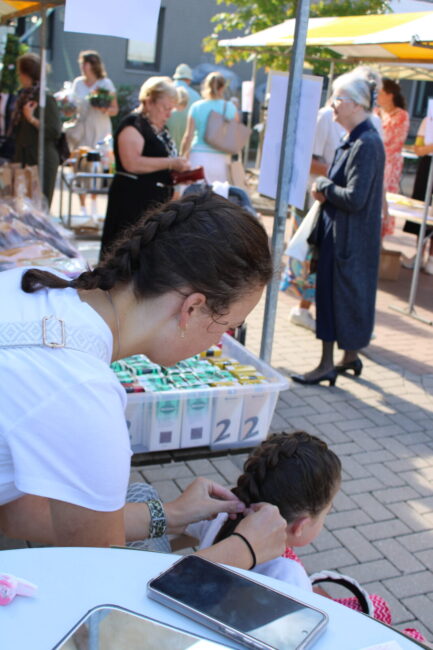 Een vrouw vlecht het haar van een kind op een markt met kraampjes. Mensen lopen rond en het is een zonnige dag.