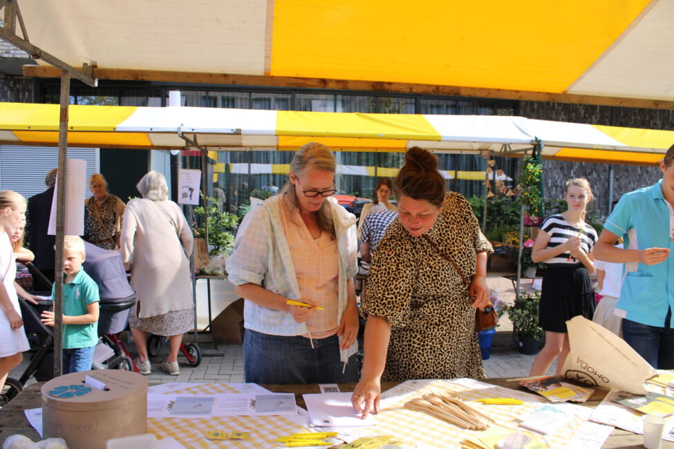 Twee vrouwen bekijken papieren op een tafel onder een geel-witte marktkraam. Mensen lopen rond op de achtergrond.