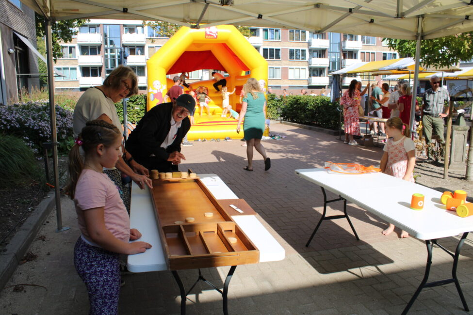 Mensen spelen sjoelbak onder een tent op een zonnige dag. Op de achtergrond is een groot, geel springkussen te zien.