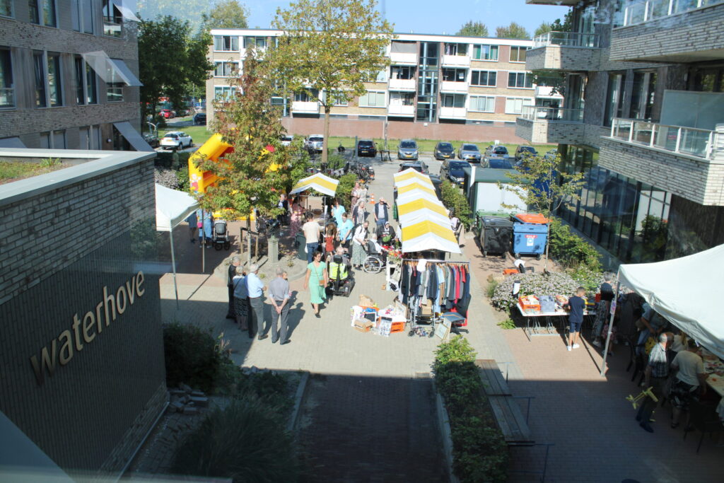 Luchtfoto van een levendige markt in een binnenplaats met kraampjes, mensen die rondlopen en een gebouw met de naam "Waterhove".