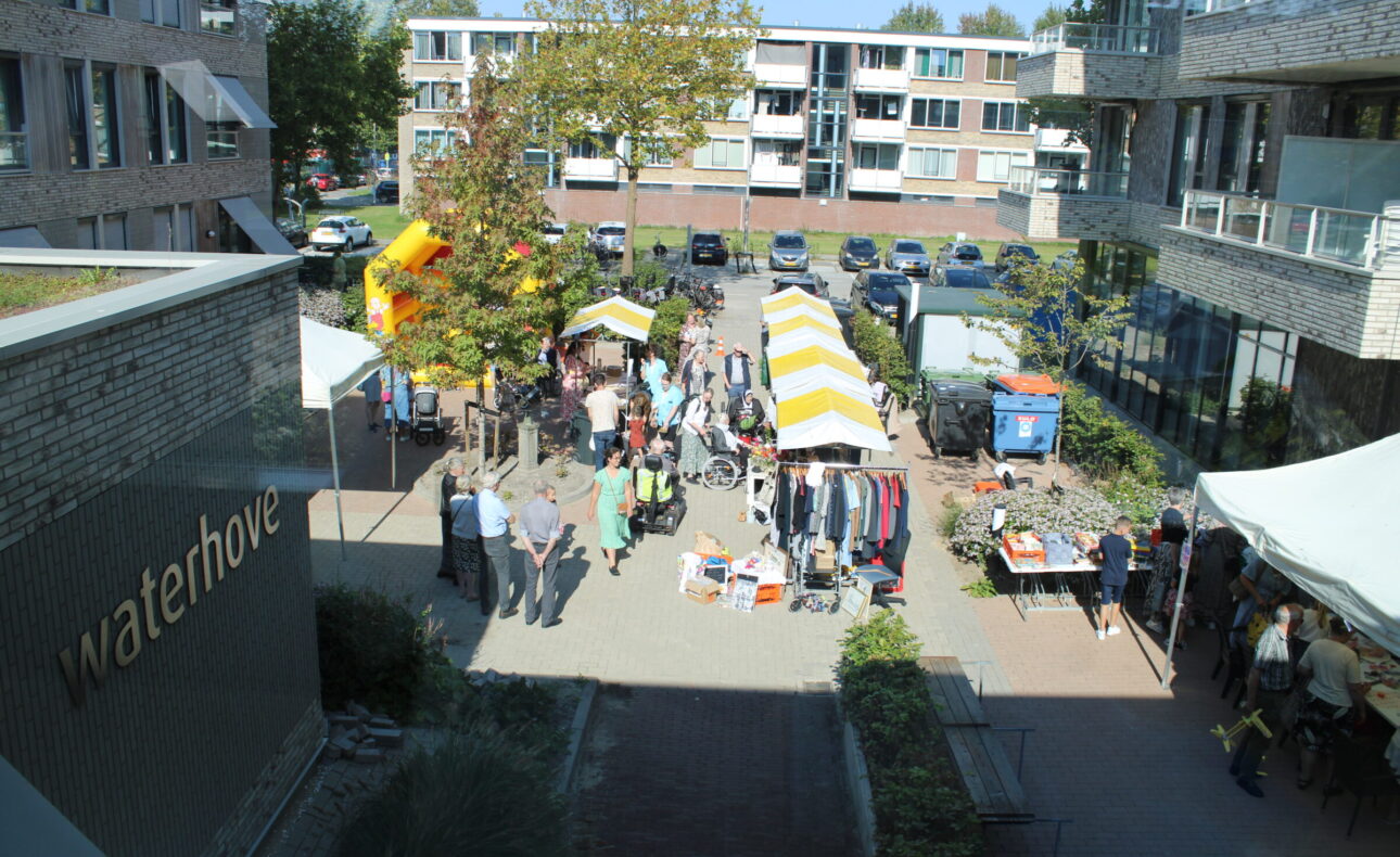 Luchtfoto van een levendige markt in een binnenplaats met kraampjes, mensen die rondlopen en een gebouw met de naam "Waterhove".