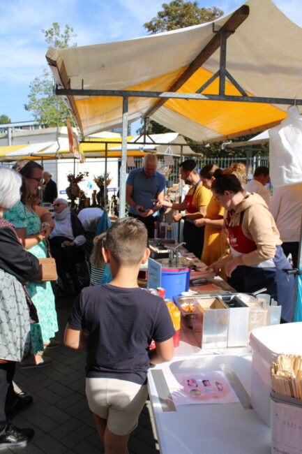 Groep mensen verzameld bij een kraam op een zonnige markt. Een jongen met een donker shirt staat op de voorgrond.