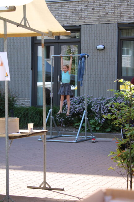 Meisje in een blauwe rok hangt aan een turnrek buiten, omringd door planten en gebouwen, op een zonnige dag.