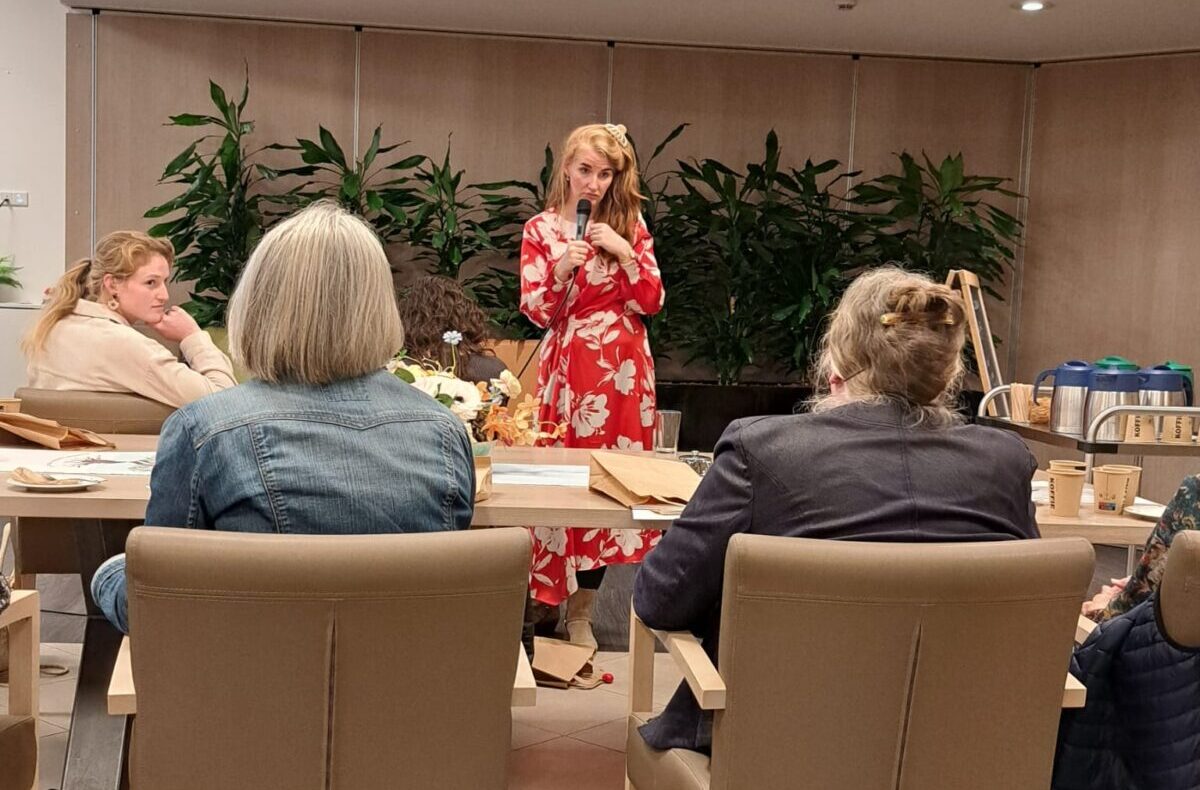 Een vrouw in een rode jurk spreekt voor een kleine groep mensen in een zaal met planten en een moderne plafondlamp.