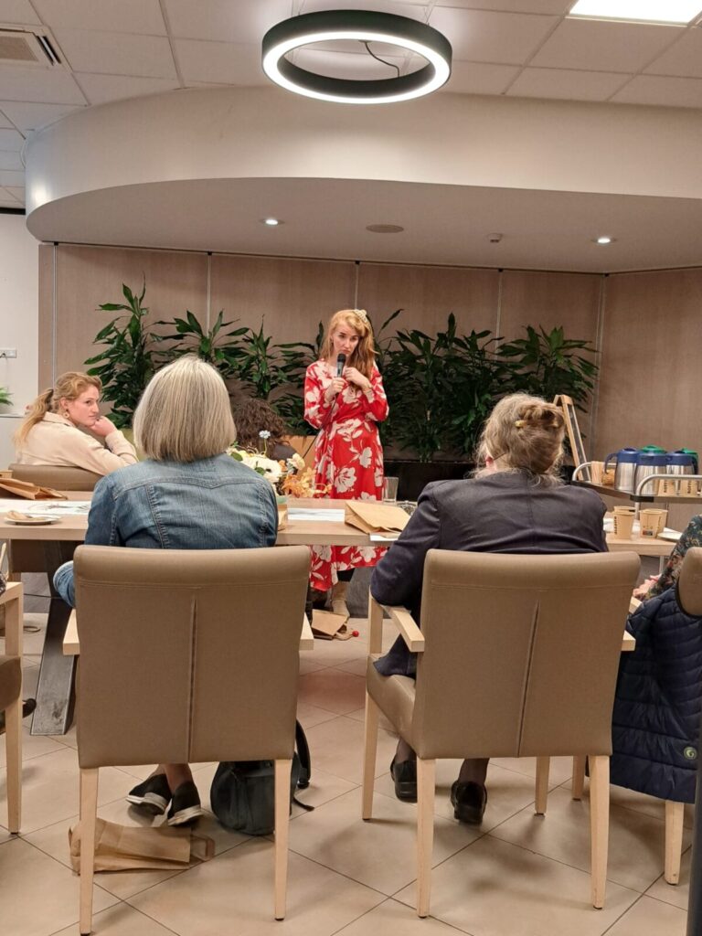 Een vrouw in een rode jurk spreekt voor een kleine groep mensen in een zaal met planten en een moderne plafondlamp.