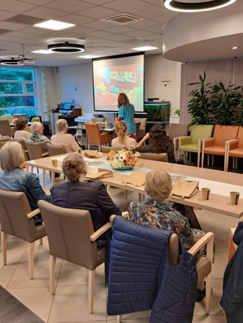 Een groep ouderen zit in een gemeenschappelijke ruimte en kijkt naar een presentatie op een groot scherm.