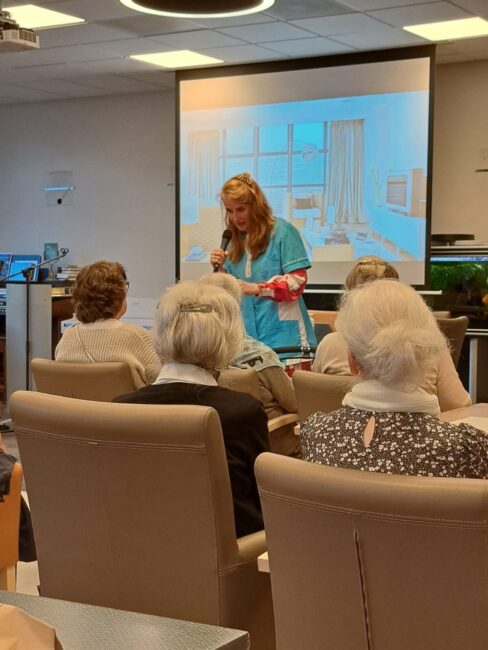 Een vrouw spreekt voor een groep ouderen in een zaal. Op de achtergrond is een projectiescherm met een afbeelding zichtbaar.