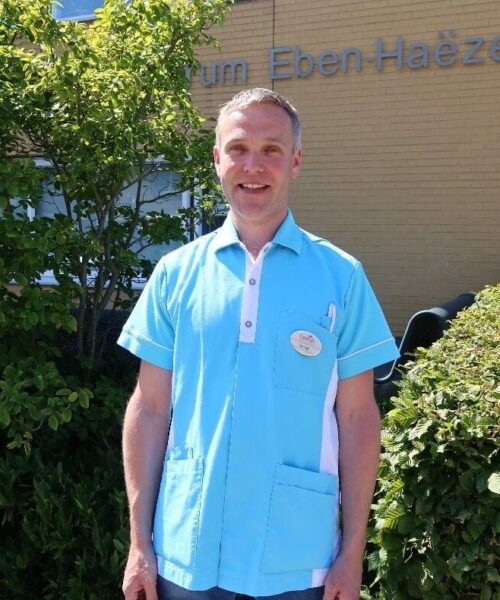 Man in blauw uniform staat buiten voor een gebouw met de naam "Eben Haëzer". Hij glimlacht en is omringd door groen.