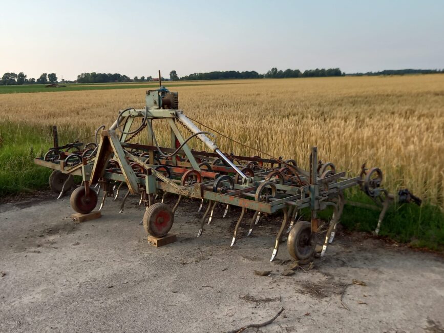 Landbouwmachine op een betonnen pad naast een korenveld onder een heldere lucht. Op de achtergrond zijn bomen zichtbaar.