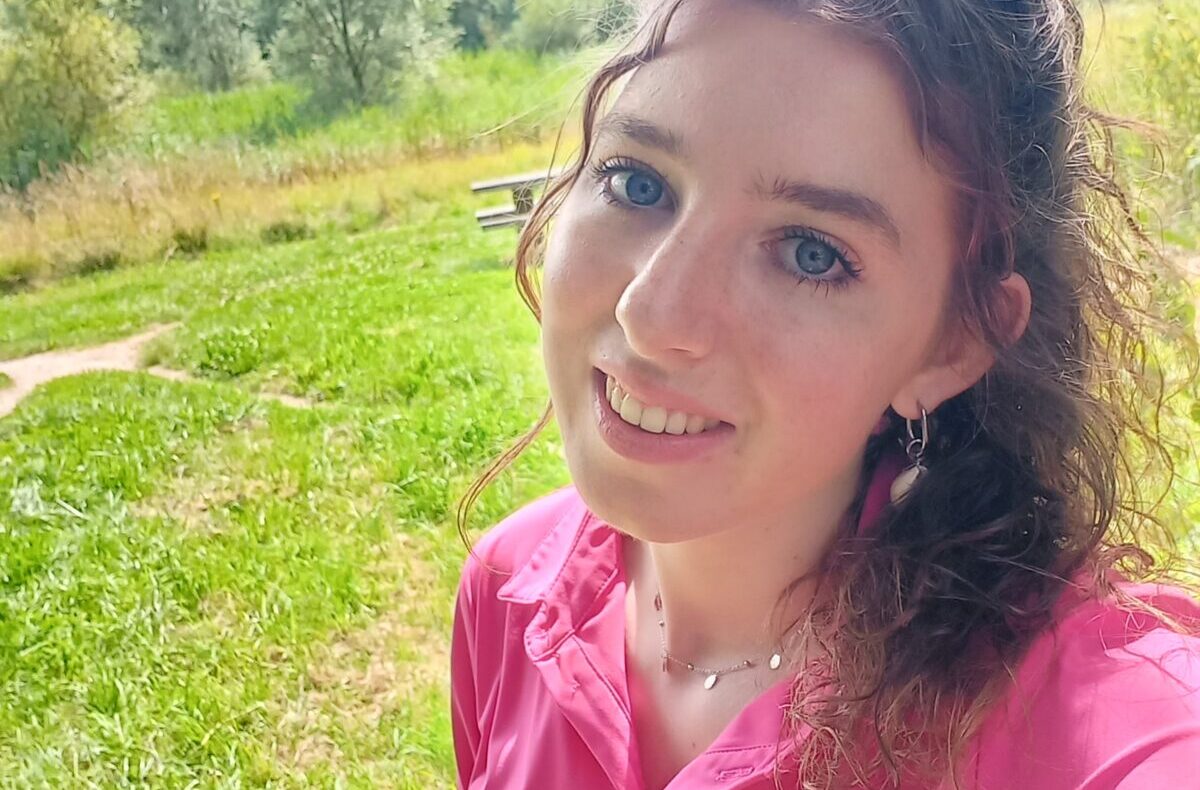 Vrouw met roze blouse glimlacht naar de camera, staand in een groene, bosrijke omgeving met bomen en gras op de achtergrond.