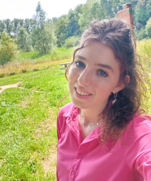 Een vrouw met krullend haar draagt een roze blouse en glimlacht naar de camera. Ze staat buiten in een groene, bosrijke omgeving.