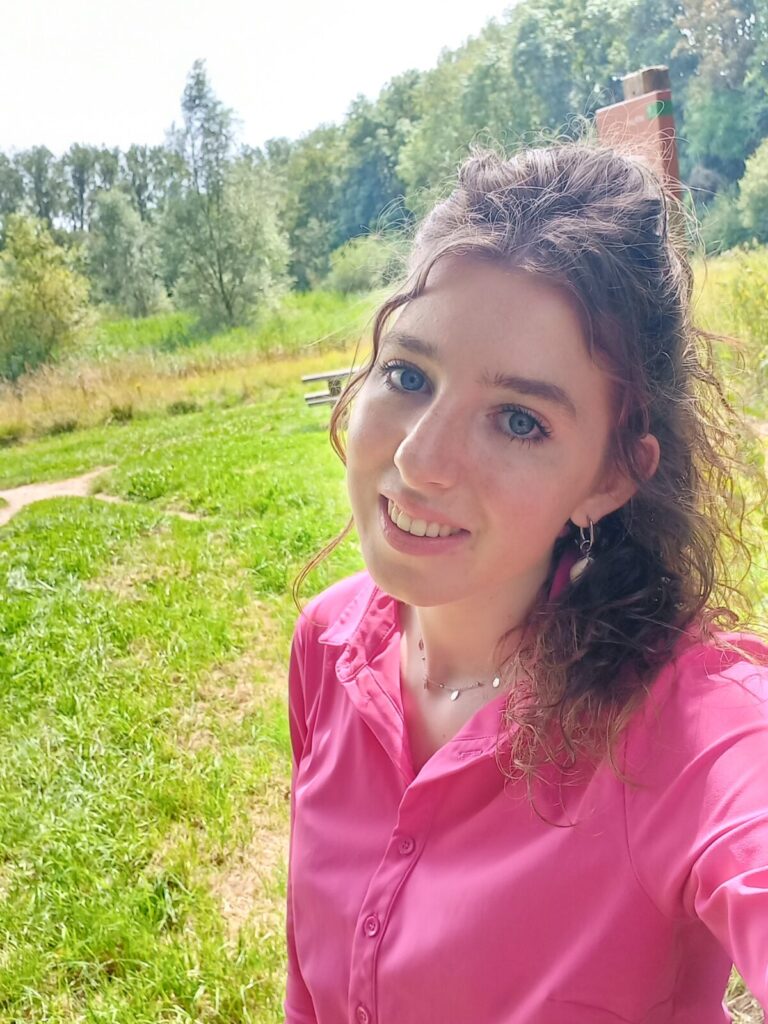 Vrouw met roze blouse glimlacht naar de camera, staand in een groene, bosrijke omgeving met bomen en gras op de achtergrond.