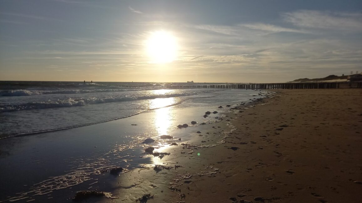 Zonsondergang aan een rustig strand met golven die het zand raken. De lucht is deels bewolkt en de horizon is zichtbaar.