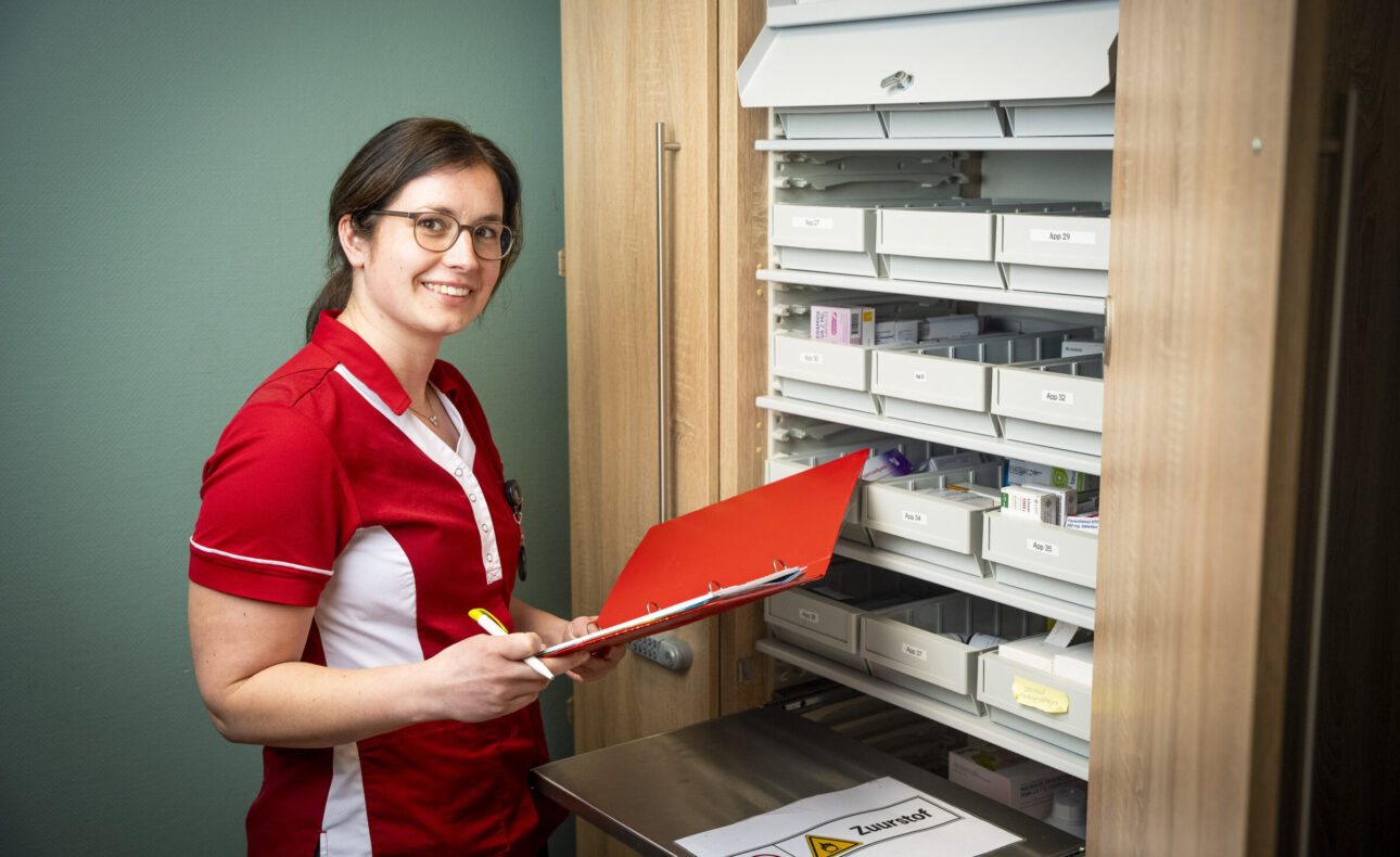 Vrouw in rood-wit uniform staat met een rode map voor een medicijnkast, glimlachend naar de camera in een zorgomgeving.