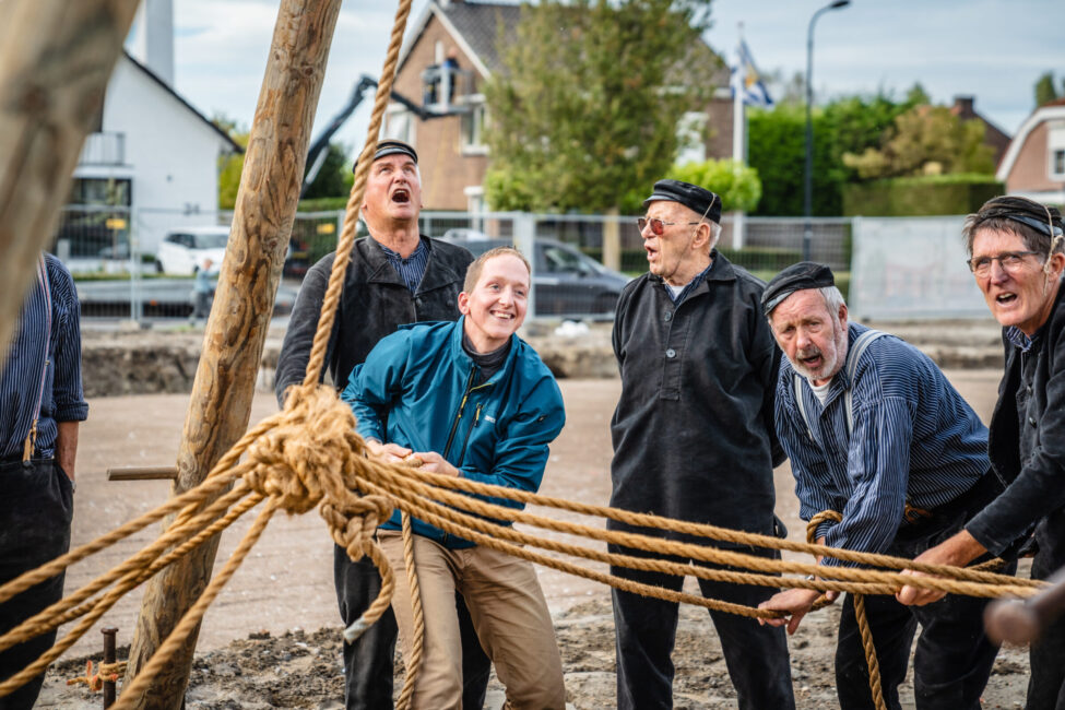 Groep mannen trekt aan touwen tijdens een buitenactiviteit. Ze lachen en dragen casual kleding. Huizen op de achtergrond.