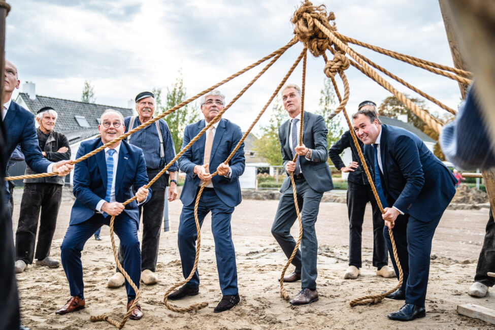 Mannen in pakken trekken aan touwen tijdens een buitenactiviteit. Ze lijken samen te werken onder een bewolkte hemel.