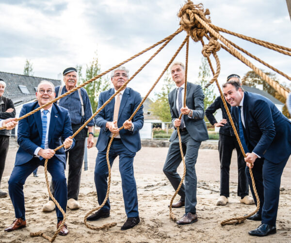 Mannen in pakken trekken aan touwen tijdens een buitenactiviteit. Ze lijken samen te werken aan een gemeenschappelijk doel.