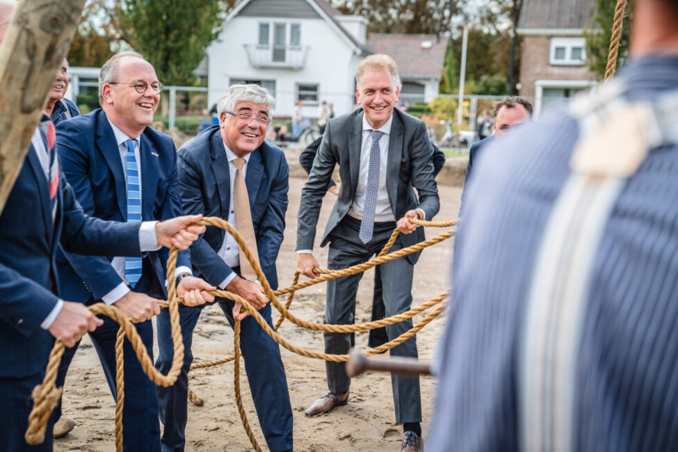 Mannen in pakken lachen terwijl ze samen aan een touw trekken tijdens een buitenactiviteit op een zonnige dag in een woonwijk.