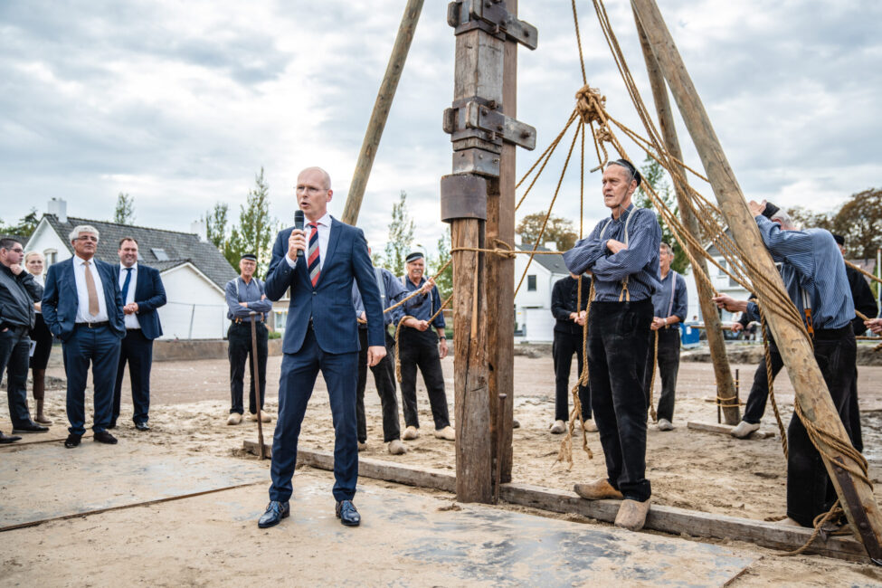 Een man in pak spreekt in een microfoon bij een bouwplaats, omringd door mensen en houten constructies op een bewolkte dag.
