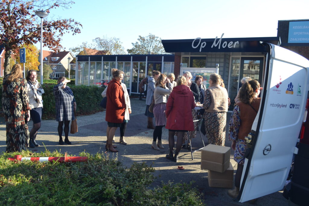 Groep mensen verzameld bij een busje voor een gebouw met de naam "Op Moer". Het is een zonnige dag met blauwe lucht.